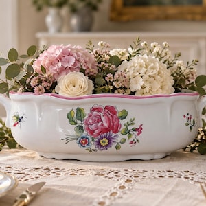 Op de afbeelding: Een witte, ovale serveerschaal van keramiek met een geschulpte rand en roze afwerking, voorzien van een centraal bloemmotief met een grote roze roos en kleinere paarse en blauwe bloemen, inclusief een bijenillustratie en twee handgrepen.