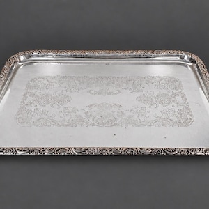 Peut inclure: Un plateau de service rectangulaire de couleur argentée avec des motifs floraux et des volutes en relief sur le bord surélevé et les poignées. La surface centrale est lisse et réfléchissante, encadrée par un rebord décoratif détaillé.