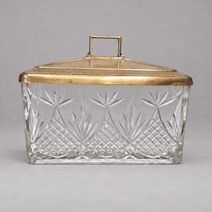 May include: A rectangular clear glass box with a cut crystal pattern featuring diamond and fan motifs. The container is topped with a metallic gold-toned lid that has a flat rectangular handle centered on the top surface.