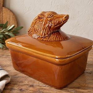 Peut inclure: Un beurrier rectangulaire en céramique marron avec une finition brillante. Le couvercle présente une tête de sanglier sculptée en trois dimensions servant de poignée. La base est un récipient simple aux parois lisses pour le beurre.