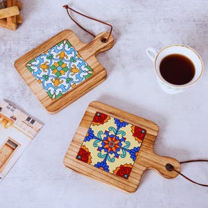 May include: Two wooden serving boards with handles and leather hanging cords. Each board features a square ceramic tile inlay with colorful geometric patterns. One tile has blue, yellow, and green motifs, while the other has red, blue, and orange designs.
