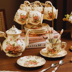 May include: A ceramic tea set featuring a cream-colored base with pink rose floral patterns and gold trim. The set includes a teapot, multiple cups and saucers, a gold metal display rack, and matching spoons with floral handles and metal bowls.