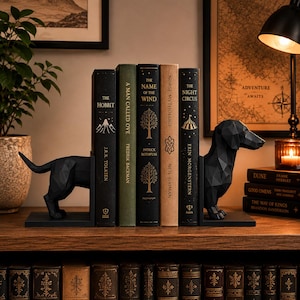 Peut inclure: Des serre-livres en forme de teckel noir géométrique soutiennent une rangée de livres. Les titres incluent "The Hobbit" et "The Night Circus". La scène est sur une étagère en bois, avec une lampe et une bougie.