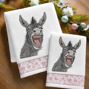 May include: Two white cotton towels featuring an embroidered gray donkey with an open mouth showing teeth and tongue. Each towel has a decorative light pink band at the bottom with a repeating floral pattern. The donkey design is detailed.
