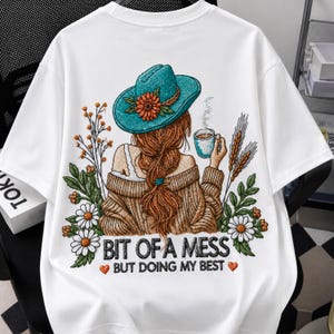 Könnte beinhalten: Ein weißes T-Shirt mit einer Grafik einer Person mit langem Haar, die einen blaugrünen Hut mit einer orangefarbenen Blume trägt und eine dampfende Tasse hält. Das Design zeigt Gänseblümchen, Weizen und den Text "BIT OF A MESS BUT DOING MY BEST".