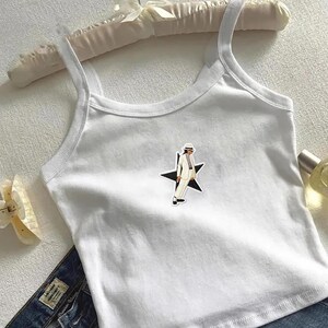 May include: A white sleeveless tank top featuring a central graphic of a figure wearing a white suit and fedora hat, positioned in front of a black star. The garment has a scoop neckline and thin shoulder straps, constructed from a plain white fabric.