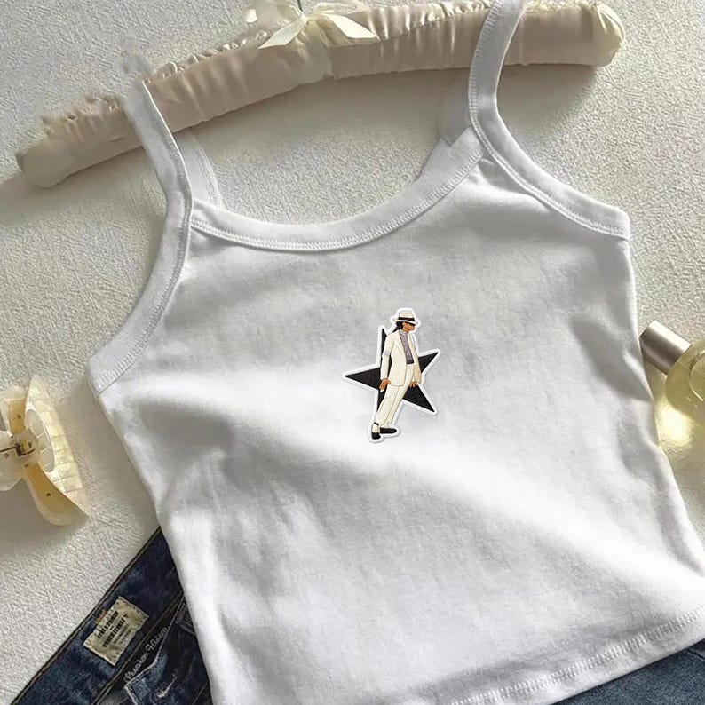 May include: A white sleeveless tank top featuring a central graphic of a figure wearing a white suit and fedora hat, positioned in front of a black star. The garment has a scoop neckline and thin shoulder straps, constructed from a plain white fabric.