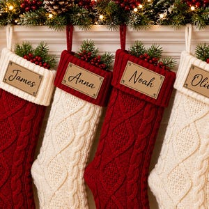 May include: Four cable-knit Christmas stockings hang from a decorated mantel. Two are red, two are cream-colored, each with a personalized name tag: James, Anna, Noah, and Oliver. The Oliver stocking has a paw print. Greenery and berries adorn the top.