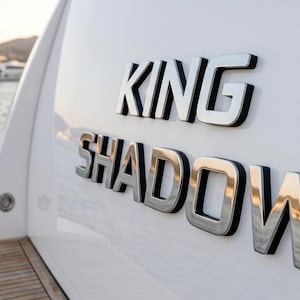 Op de afbeelding: De witte buitenkant van een jacht is voorzien van de naam KING SHADOW in grote, reflecterende, verchroomde 3D-letters. De letters zijn gemonteerd op de gebogen witte romp, geplaatst boven een houten dekgedeelte nabij het wateroppervlak.
