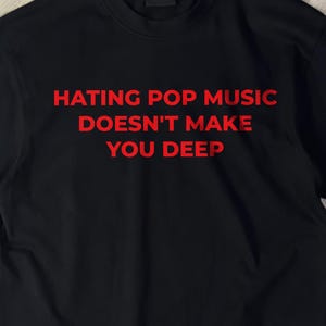 May include: A black short-sleeved t-shirt featuring the red text "HATING POP MUSIC DOESN'T MAKE YOU DEEP" printed in a bold, sans-serif font across the center of the chest. The garment has a classic crew neckline and a straight hem.