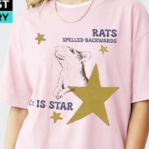 May include: A light pink short-sleeve t-shirt featuring a graphic of a rat and several gold stars. The text reads RATS SPELLED BACKWARDS IS STAR. The shirt has a crew neckline and is layered over a white undershirt, paired with black shorts.