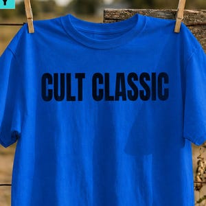 Pode incluir: Uma t-shirt de manga curta azul brilhante com o texto CULT CLASSIC impresso em letras maiúsculas pretas em negrito no peito. A peça tem um decote redondo padrão e está pendurada por um prendedor de roupa de madeira numa cerca de arame.