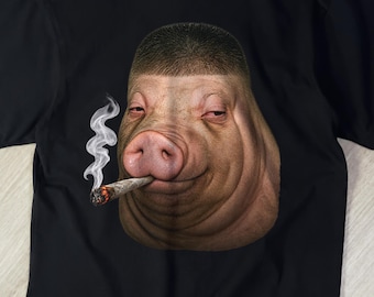 May include: A black short-sleeved t-shirt featuring a graphic print of a pig with a bowl-cut hairstyle. The pig has a relaxed expression and is holding a lit cigarette in its mouth, with a trail of white smoke rising from the glowing ember.