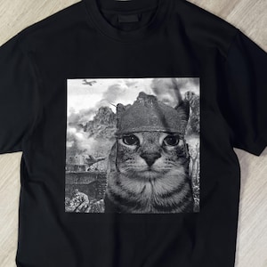 Peut inclure: Un t-shirt noir à manches courtes avec une impression graphique centrale en noir et blanc. L'image représente un chat portant un casque de style militaire, sur un fond de champ de bataille avec des montagnes, de la fumée et un petit avion.