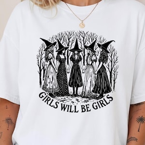 May include: A white short-sleeve t-shirt featuring a black graphic print of five witches in pointed hats holding hands in a forest setting. Below the illustration, the text reads GIRLS WILL BE GIRLS in a bold, black, capitalized sans-serif font.