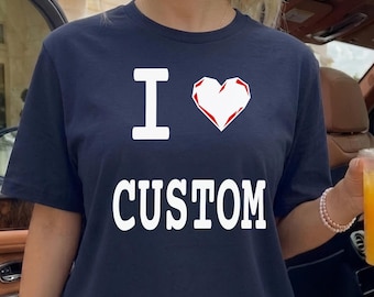 May include: A navy blue short-sleeved t-shirt featuring the white text I followed by a white heart graphic and the word CUSTOM in white capital letters. The shirt has a crew neckline and is worn with a white pleated skirt, creating a casual look.