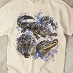 Pode incluir: Uma t-shirt de manga curta cor de creme com uma impressão gráfica de três crocodilos e uma lua cheia. O design inclui um crocodilo central numa pose de caminhada, um grande plano de uma cabeça de crocodilo e um perfil lateral.