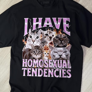 Peut inclure: Un t-shirt noir à manches courtes avec une impression graphique de cinq chats entourés de flammes. Le texte "I HAVE HOMOSEXUAL TENDENCIES" est imprimé dans une police violette claire et usée au-dessus et au-dessous des images de chats.