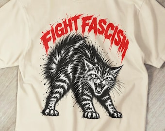 May include: A cream-colored short-sleeve t-shirt featuring a black and white illustration of an arched-back hissing cat. Above the cat, the text "FIGHT FASCISM" is printed in a bold, distressed red font with a dripping paint effect.