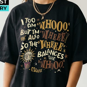 Può includere: Una t-shirt nera a maniche corte con una stampa grafica in testo dorato e bianco che recita: I TOO AM WHOOO BUT I'M ALSO SO THE WHEEE WHEEE BALANCES THE WHOOO ESQUIE. Il design include una piccola icona simile a un sole con un volto e delle stelle.