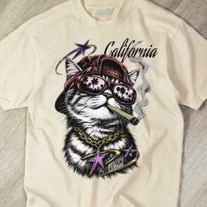 Può includere: Una t-shirt color crema con la grafica di un gatto che indossa un berretto da baseball rosso e occhiali da sole con riflessi di palme. Il gatto ha una catena d'oro, un sigaro ed è circondato da stelle viola e dalla scritta California in corsivo.