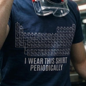 Op de afbeelding: Een marineblauw t-shirt met korte mouwen voorzien van een witte afbeelding van het periodiek systeem der elementen en de tekst I WEAR THIS SHIRT PERIODICALLY. Een doorzichtige veiligheidsbril met een rode band rust bij de hals.