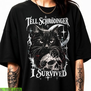 Puede incluir: Una camiseta negra de manga corta con un estampado gráfico blanco de un gato apoyando su pata sobre una calavera. El texto sobre el gato dice "TELL SCHRÖDINGER" y el texto debajo de la calavera dice "I SURVIVED". El diseño incluye estrellas y una luna creciente.