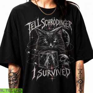 Peut inclure: Un t-shirt noir à manches courtes avec une impression graphique représentant un chat, un crâne et un cube. Le texte "TELL SCHRODINGER I SURVIVED" est écrit dans une police stylisée. Le motif inclut des lignes orbitales, des étoiles et une lune.