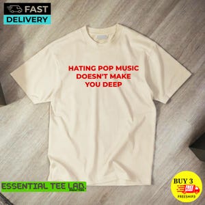 May include: A cream-colored short-sleeve t-shirt featuring the red text "HATING POP MUSIC DOESN'T MAKE YOU DEEP" printed in the center of the chest. The garment has a classic crew neckline and a standard fit with short sleeves.