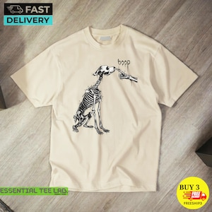 May include: A cream-coloured short-sleeve t-shirt featuring a black ink-style graphic of a dog skeleton being poked on the nose by a skeletal finger. Above the dog, the word boop is written in a stylised, dripping black font.
