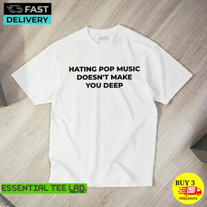 May include: A white short-sleeved t-shirt featuring the black text HATING POP MUSIC DOESN'T MAKE YOU DEEP printed in the center of the chest. The garment has a crew neckline and a standard fit, presented on a light-colored wooden surface.