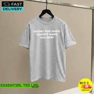 May include: A heather gray short-sleeve crew neck t-shirt featuring the white text HATING POP MUSIC DOESN'T MAKE YOU DEEP printed on the front. The garment is displayed on a black hanger against a neutral background.