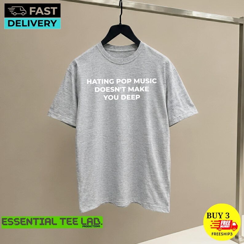 May include: A heather gray short-sleeve crew neck t-shirt featuring the white text HATING POP MUSIC DOESN'T MAKE YOU DEEP printed on the front. The garment is displayed on a black hanger against a neutral background.