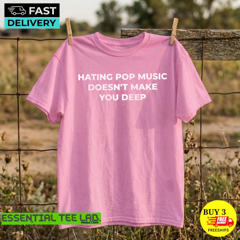 May include: A light pink short-sleeved t-shirt featuring the white text "HATING POP MUSIC DOESN'T MAKE YOU DEEP" printed across the center of the chest. The garment has a crew neckline and a standard fit, displayed hanging on a wooden clothesline.