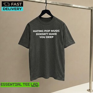 May include: A dark charcoal gray short-sleeved t-shirt featuring the white text "HATING POP MUSIC DOESN'T MAKE YOU DEEP" printed across the chest. The shirt is displayed on a black hanger against a neutral background.