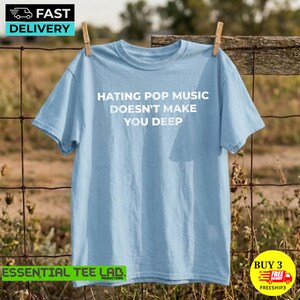 May include: A light blue short-sleeve t-shirt featuring the white text "HATING POP MUSIC DOESN'T MAKE YOU DEEP" printed in the center of the chest. The garment has a crew neckline and a standard fit, displayed hanging from a wooden clothespin.