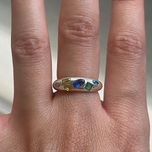 May include: A silver-toned band ring featuring a smooth, rounded surface embedded with multiple small, multi-colored gemstones in various shapes, including blue, green, and yellow stones arranged across the front of the metal band.