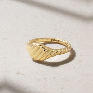 Puede incluir: Un anillo de oro amarillo pulido con una banda trenzada similar a una cuerda que se ensancha en una cúpula acanalada estilo croissant en la parte superior. El interior de la banda tiene grabado el texto 750, indicando la pureza del oro.