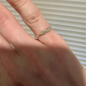 May include: A thin gold band ring featuring a curved, slightly tapered setting at the top. The setting is embedded with five small, clear, round-cut stones arranged in a row, creating a delicate and refined appearance on the finger.