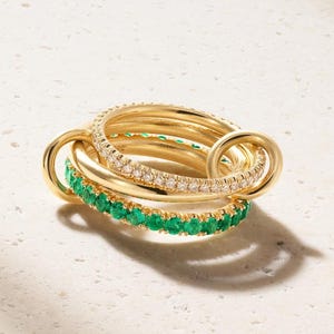 Puede incluir: Un conjunto de anillos entrelazados de oro que consta de tres bandas distintas. Una banda es de oro pulido liso, otra está engastada con piedras blancas transparentes y la tercera está engastada con piedras verdes vibrantes. Las bandas están unidas.