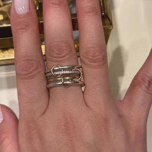 Könnte beinhalten: Ein mehrreihiger Ring aus einer Kombination von silber- und goldfarbenen Metallbändern. Das Design umfasst ein mittleres Band mit kleinen klaren Steinen, ein gedrehtes goldfarbenes Drahtband und glatte, ineinandergreifende kreisförmige Elemente.