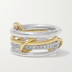 Op de afbeelding: Een ring met meerdere banden, bestaande uit een combinatie van gepolijste zilverkleurige en goudkleurige metalen banden. Een van de middelste banden is ingelegd met een rij kleine, heldere, rondgeslepen stenen, terwijl de andere banden glad en in elkaar verstrengeld zijn.