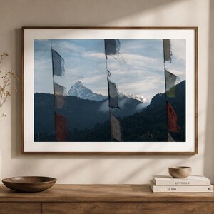 Könnte beinhalten: Gerahmte Landschaftsfotografie mit einem schneebedeckten Berggipfel hinter bunten Gebetsfahnen. Die Fahnen befinden sich im Vordergrund, mit einer Bergkette und einem bewölkten Himmel im Hintergrund. Das Foto ist in einem braunen Holzrahmen.