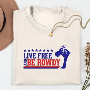 Könnte beinhalten: Cremefarbenes T-Shirt mit dem Text "LIVE FREE AND BE ROWDY" in Blau und Rot. Das Design beinhaltet eine stilisierte Figur und Sterne. Eine goldene Halskette mit Sternanhänger und ein brauner Hut sind ebenfalls zu sehen.