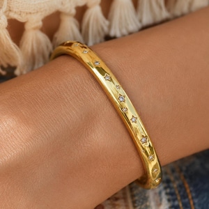 May include: A polished gold bangle bracelet featuring a series of small, evenly spaced star-shaped cutouts along the center. Each star cutout contains a single, clear, sparkling gemstone set in the middle, creating a celestial-inspired design.