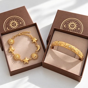 May include: A gold chain bracelet featuring sun, star, and crescent moon charms, alongside a gold cuff bracelet engraved with sun, moon, and star motifs, both presented in brown boxes with a gold sun and moon logo printed on the lids.