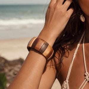 May include: A wide stretch bracelet composed of alternating rectangular wooden segments in varying shades of light tan, medium brown, and dark brown. The bracelet features a smooth, polished finish and is constructed with an elastic band for flexibility.
