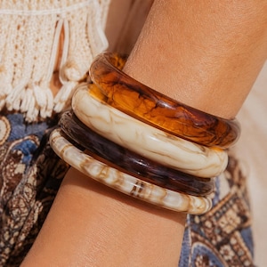 May include: A set of four circular bangle bracelets featuring a smooth, polished finish. The stack includes one translucent amber-coloured bangle, one opaque cream-coloured bangle with marble-like veining, one dark brown bangle, and one light beige bangle.
