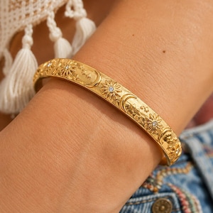 May include: A gold-toned bangle bracelet featuring a repeating celestial motif of embossed suns, crescent moons, and small stars. Several of the sun designs are accented with a small, clear, round-cut stone set in the center of each sun.