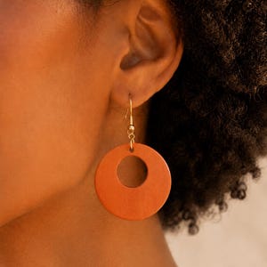 Könnte beinhalten: Ein runder Ohrring aus gebranntem orangefarbenem Holz mit einem kreisförmigen Ausschnitt in der Mitte. Der Ohrring hängt an einem dünnen, goldfarbenen Metallhaken, der mit einem kleinen Verbindungsring am oberen Teil der Holzscheibe befestigt ist.